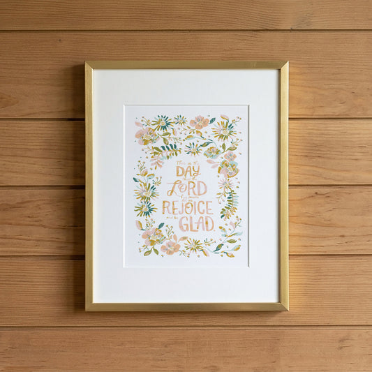 A framed art print featuring the hand-lettered quote 'DAY AND LORD AND REJOICE GLAD' surrounded by a watercolor floral wreath, displayed on a light wood plank wall.